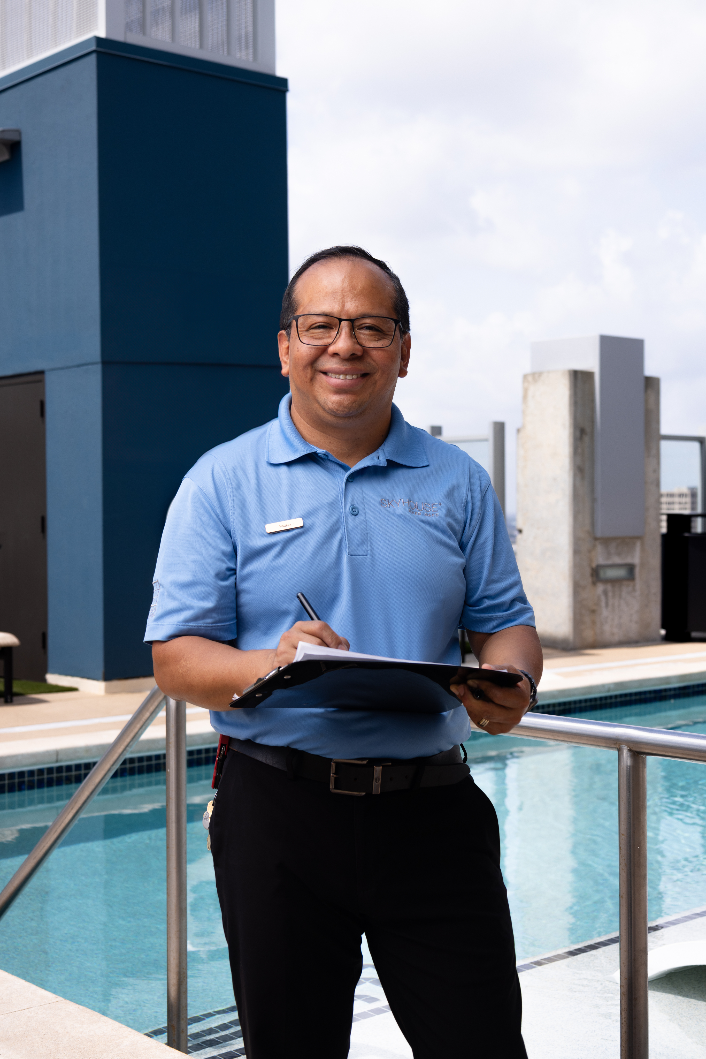 A smiling man in a blue polo shirt holds a clipboard by a pool, with a modern building in the background.