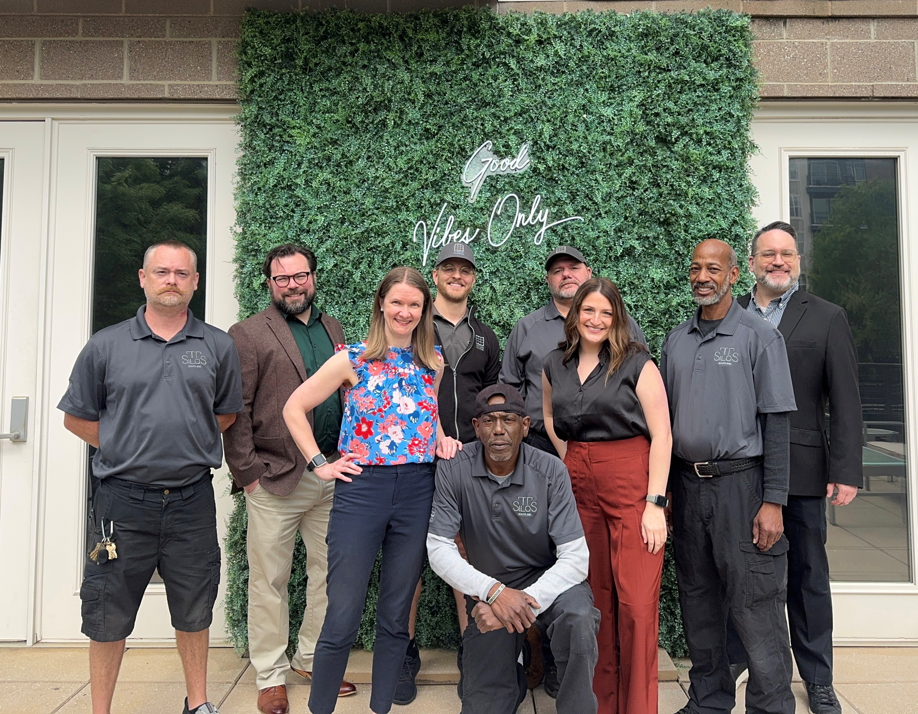 A group of nine people poses in front of a green wall with the text "Good Vibes Only." They are dressed in casual and business attire.