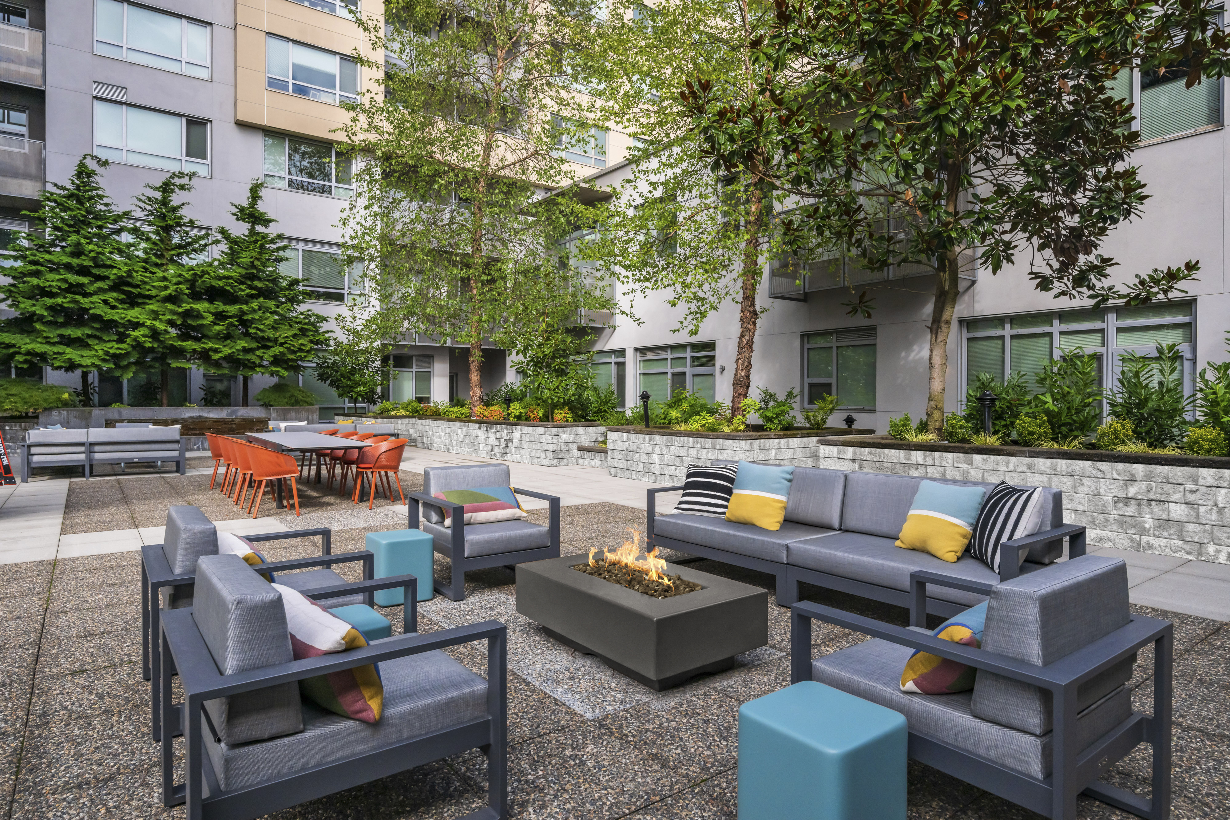 A modern outdoor lounge area featuring gray seating, colorful cushions, a fire pit, and a dining table surrounded by greenery.