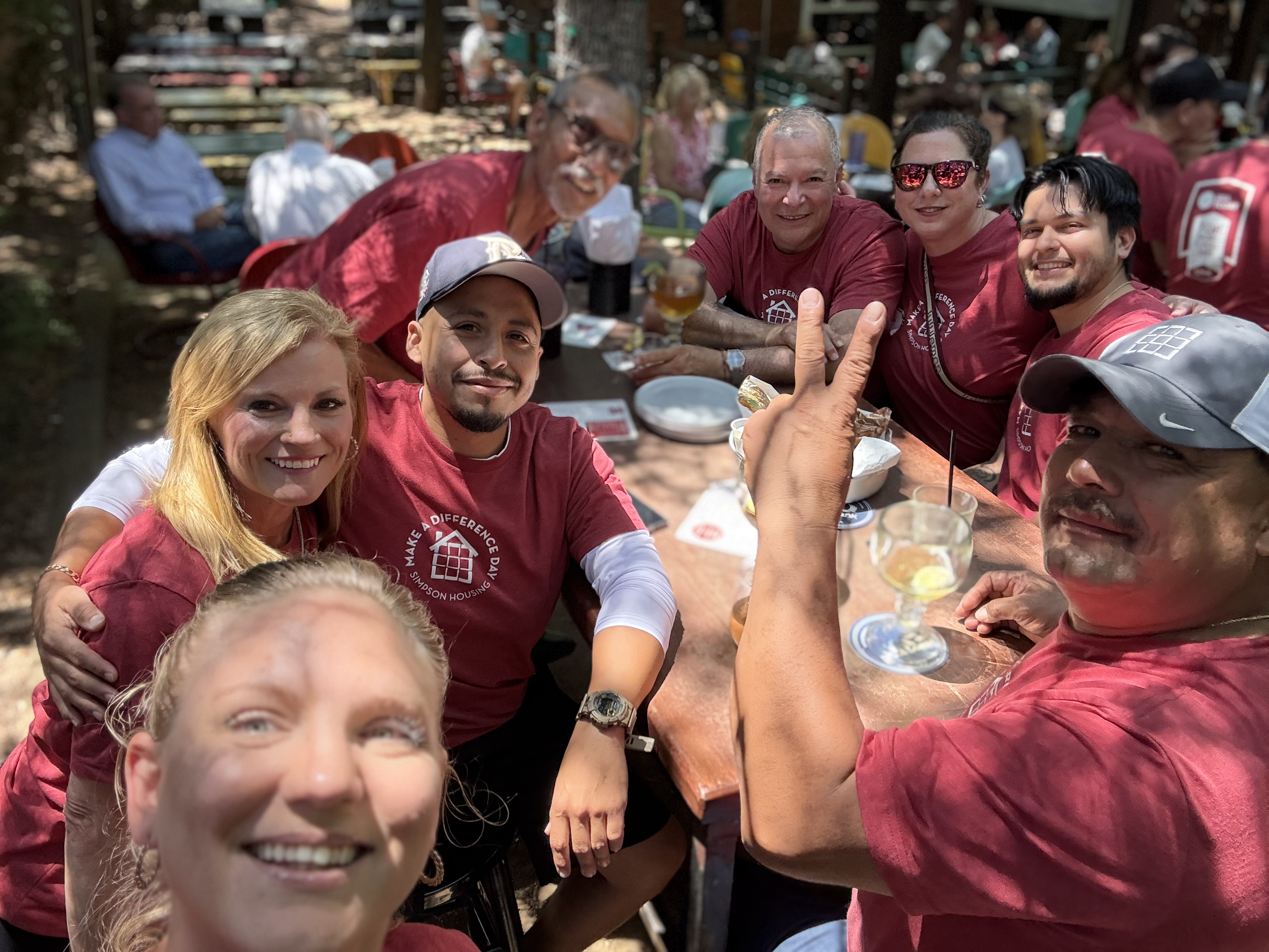 A group of eight people in red shirts smiling and posing at a table outdoors, enjoying drinks and food in a lively setting.
